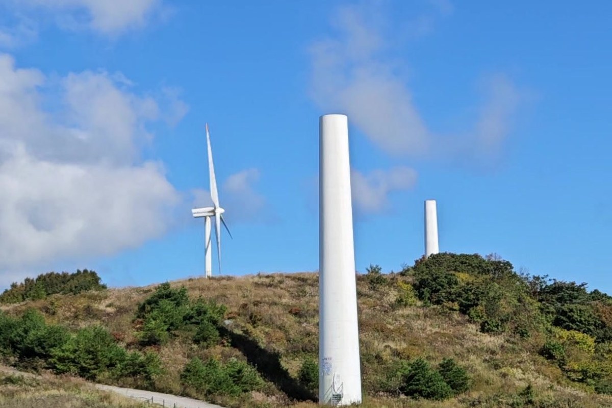 A tall white wind turbine tower beside a narrow path on a grassy ridge at Sky Ranch, with more turbines behind under a blue sky