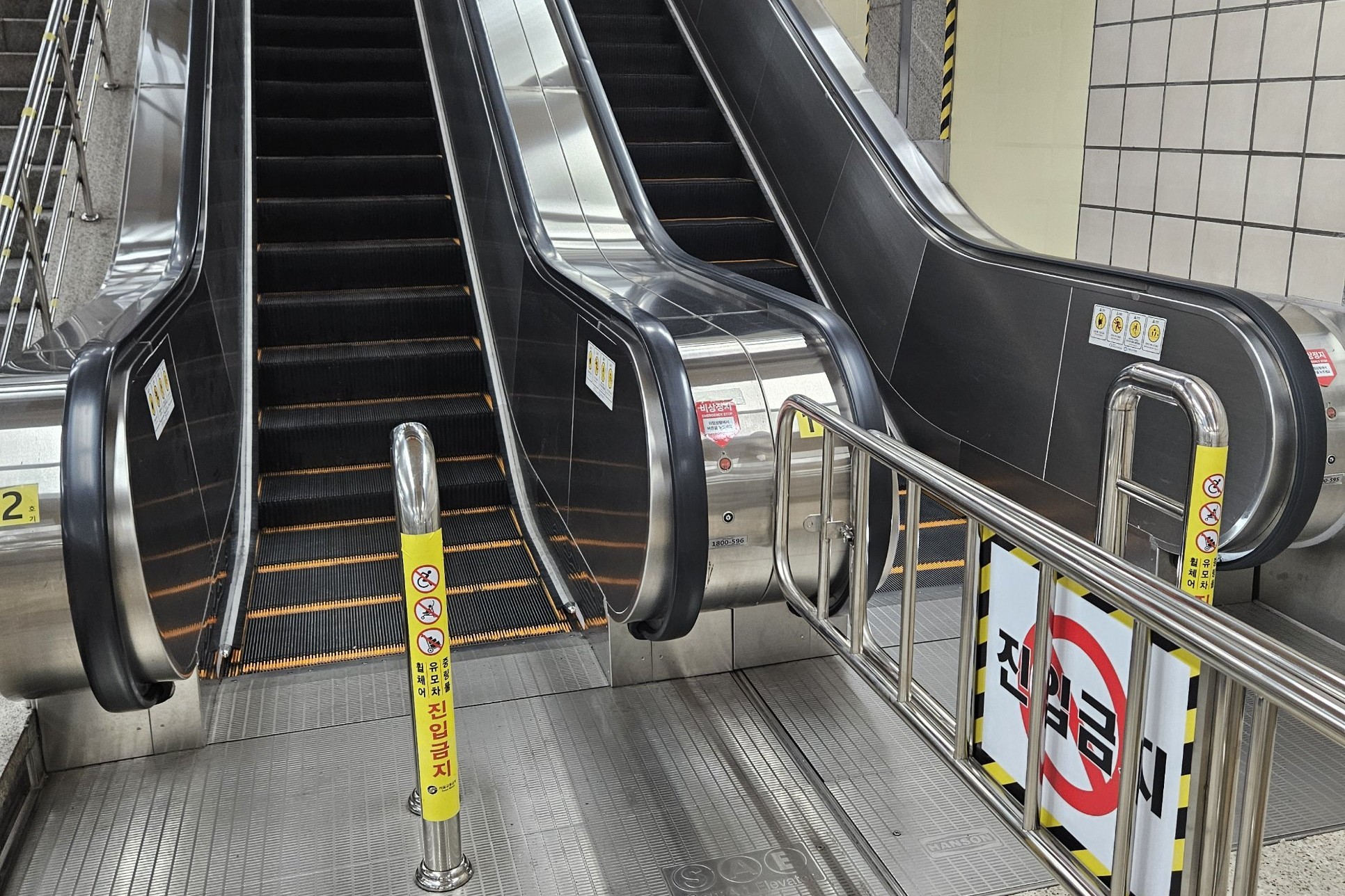 Escalators at a Seoul subway transfer with a yellow post showing no wheelchairs, strollers or carts, and a 진입금지 (no entry) barrier