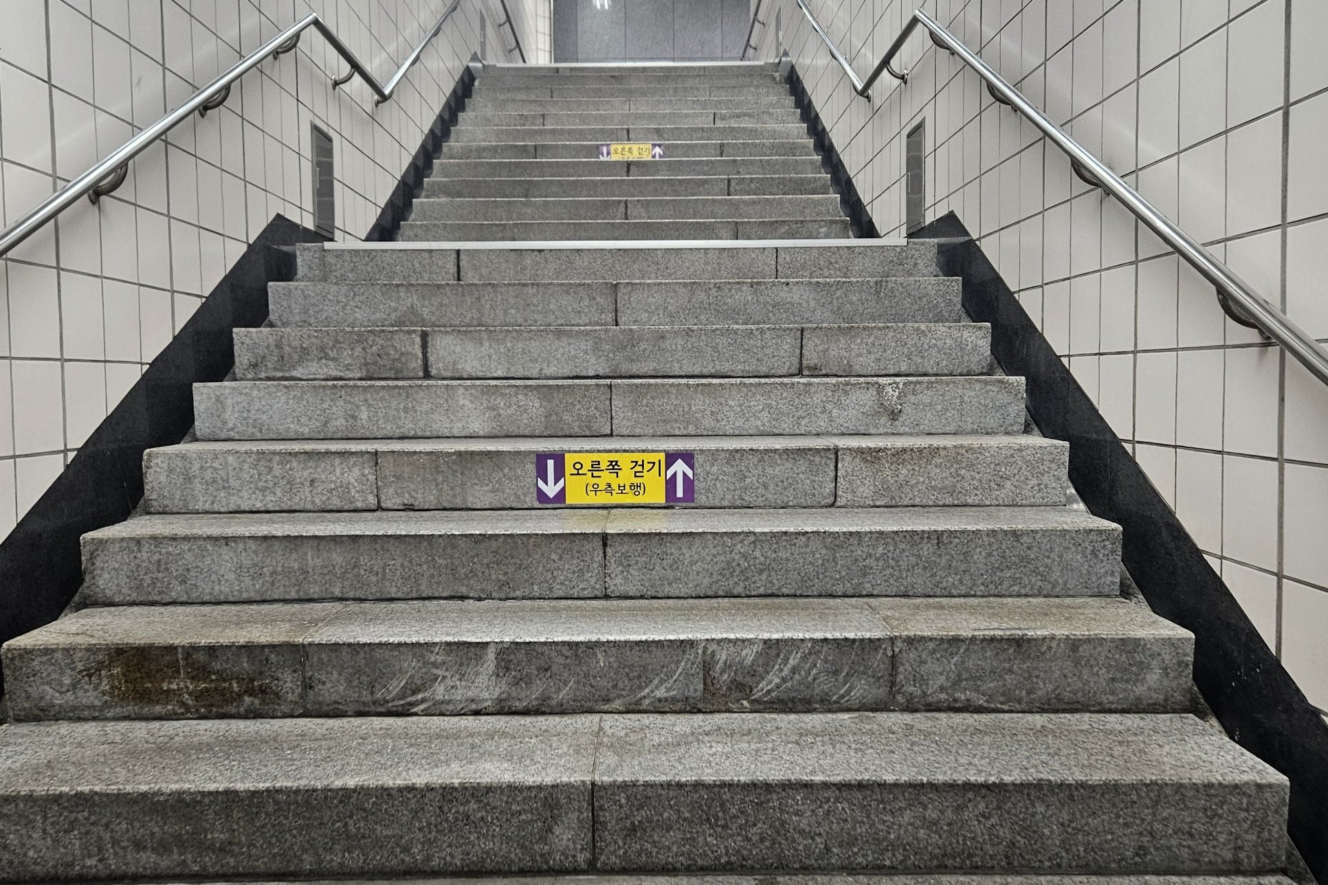 A long flight of granite stairs inside a Seoul subway station with a yellow sticker reading 오른쪽 걸기 (우측보행), keep right