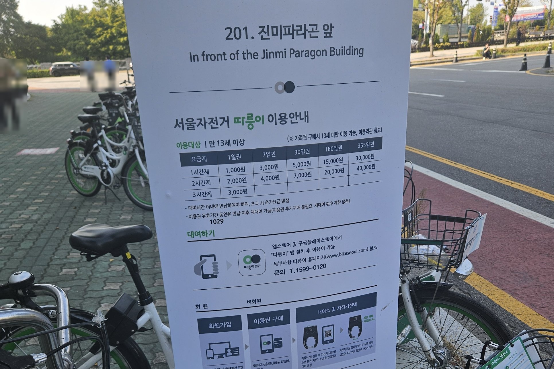 Ttareungyi station guide board 201, In front of the Jinmi Paragon Building, with a price table: 1-day pass 1,000 won for 1 hour, 2,000 for 2 hours, 3,000 for 3 hours; ages 13 and over