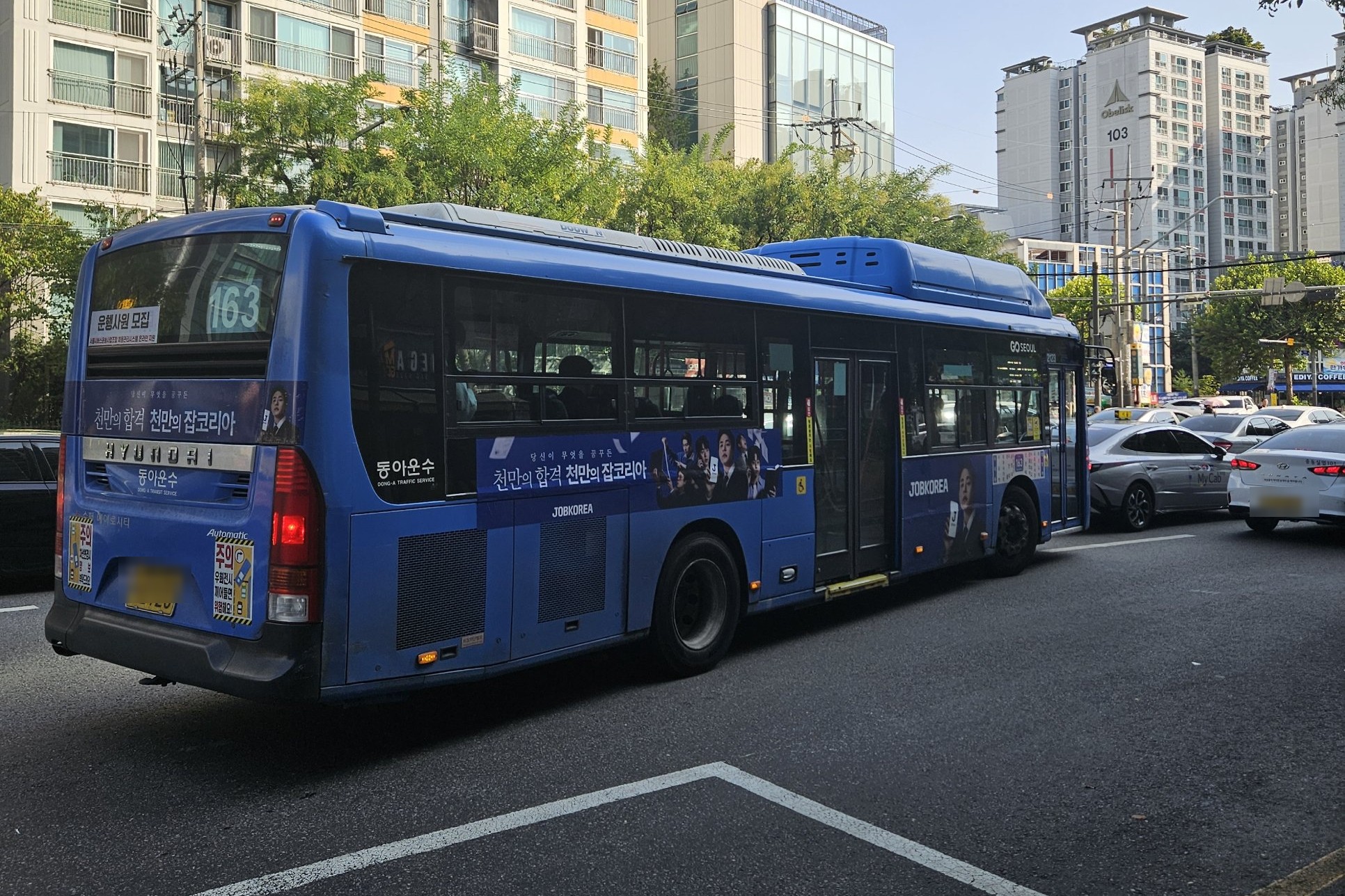 A blue Seoul trunk bus, route 163, pulling away from a stop on a Seoul street, Sept 2026