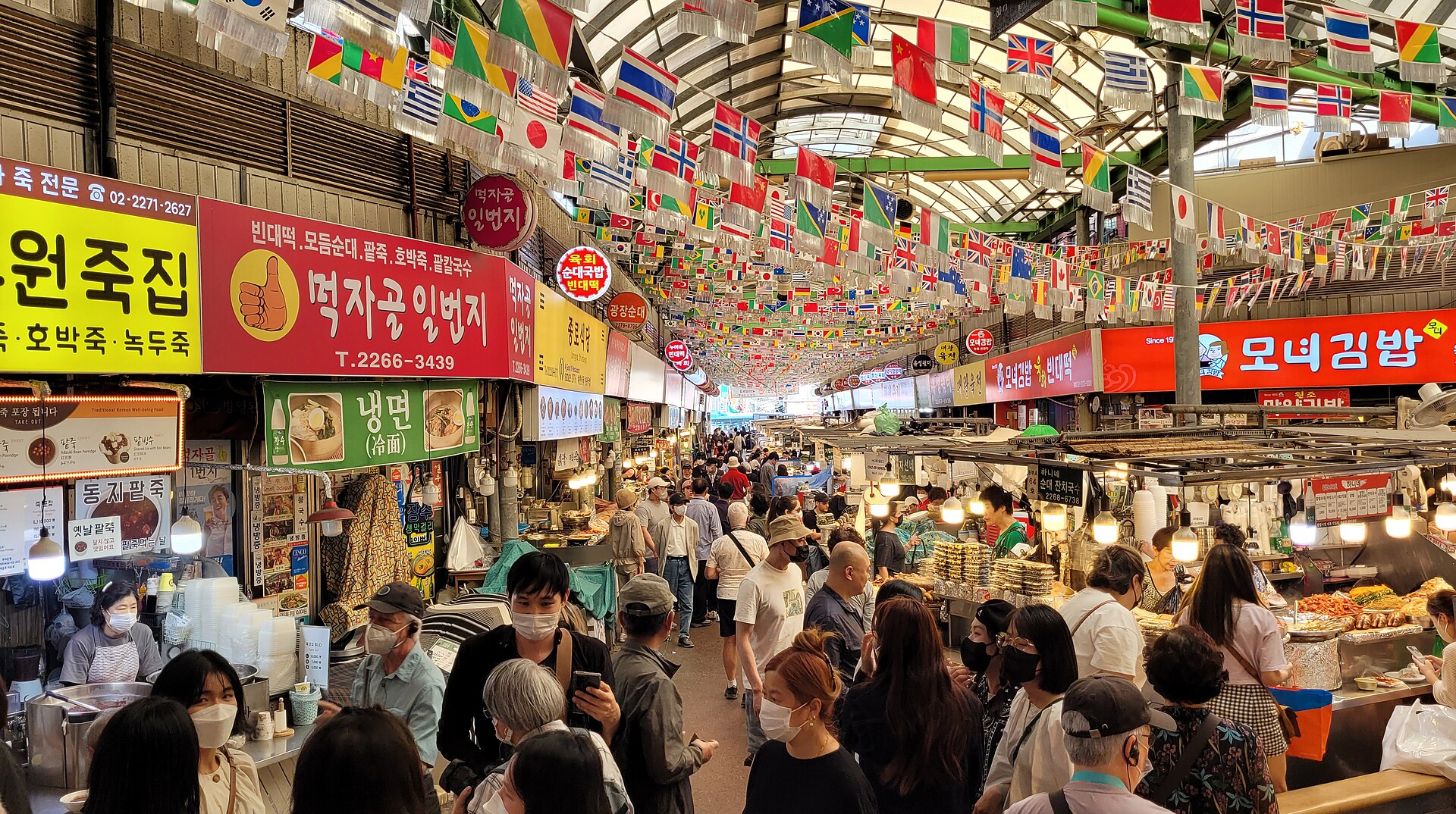 Busy covered food alley in Gwangjang Market, Seoul, with stall signs in Korean and flags hanging from the roof