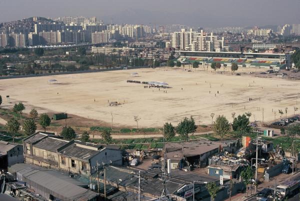 Bare dirt field of the former Ttukseom racecourse in 1999, with factory roofs in front and apartment blocks behind