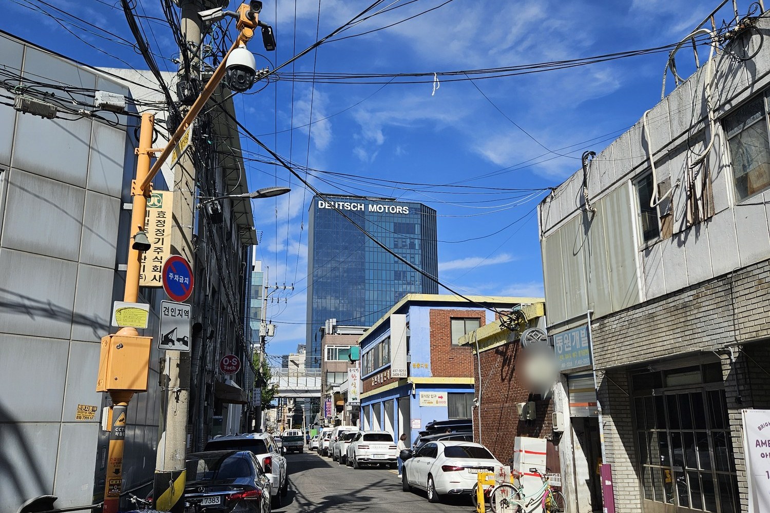 A narrow Seongsu side street with low workshop buildings, overhead power lines and a glass office tower at the end