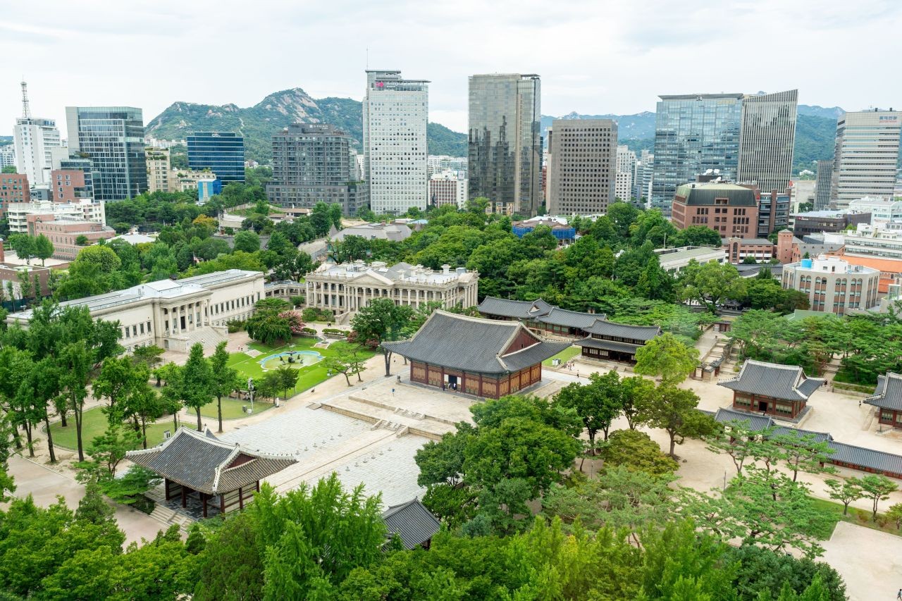 Deoksugung Palace seen from above, with palace roofs and trees in front of downtown Seoul office towers and mountains