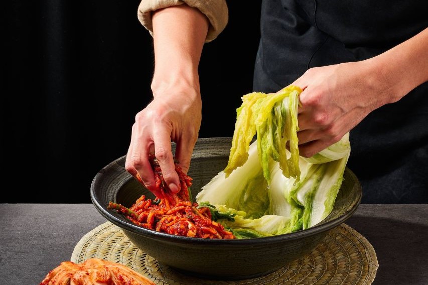 Hands spreading red seasoning between the leaves of salted cabbage in a dark bowl