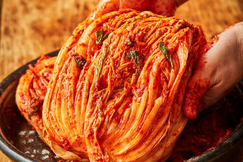 Hands holding a half head of salted cabbage covered in red kimchi seasoning over a bowl