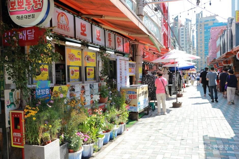 A sunny lane at Haeundae Market in Busan lined with food shops, awnings, flower pots and signs, with shoppers walking in the distance