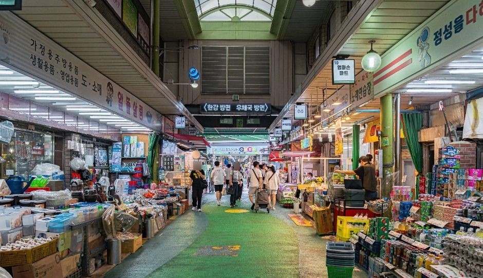 The covered arcade of Mangwon Market in Seoul with a green walkway, shops selling kitchenware and groceries on both sides, and shoppers walking away from the camera