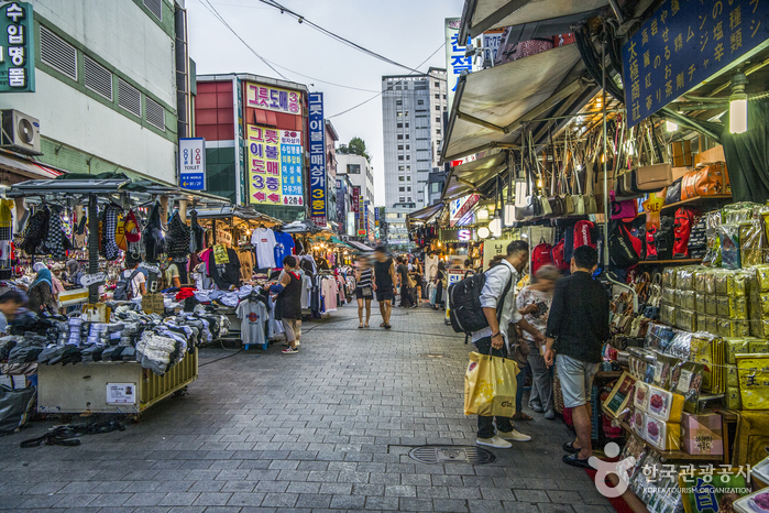 A crowded lane in Namdaemun Market lined with clothing stalls and signs, with visitors carrying shopping bags