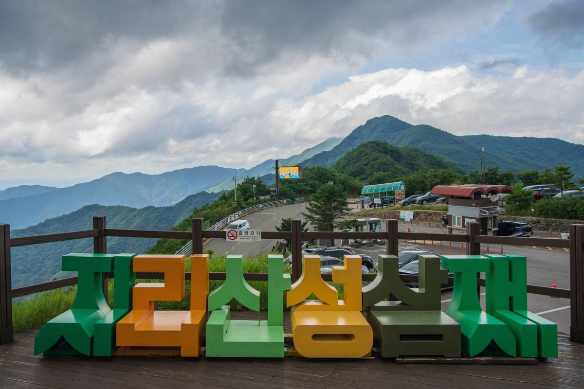 Large colourful letters spelling Jirisan Seongsamjae at a viewpoint with a car park and ridges behind under a cloudy sky