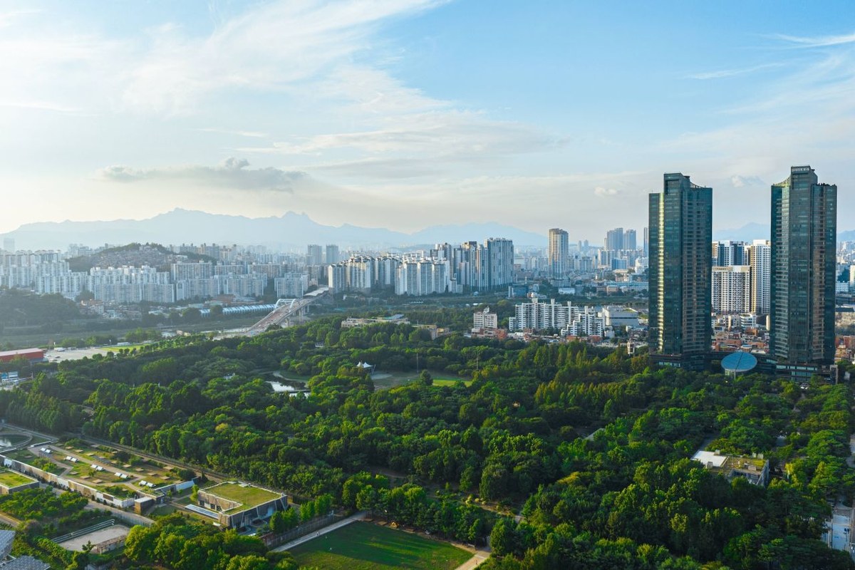 Aerial view of Seoul Forest with green trees, a water-treatment park and the Han River side with high-rise towers on a clear day