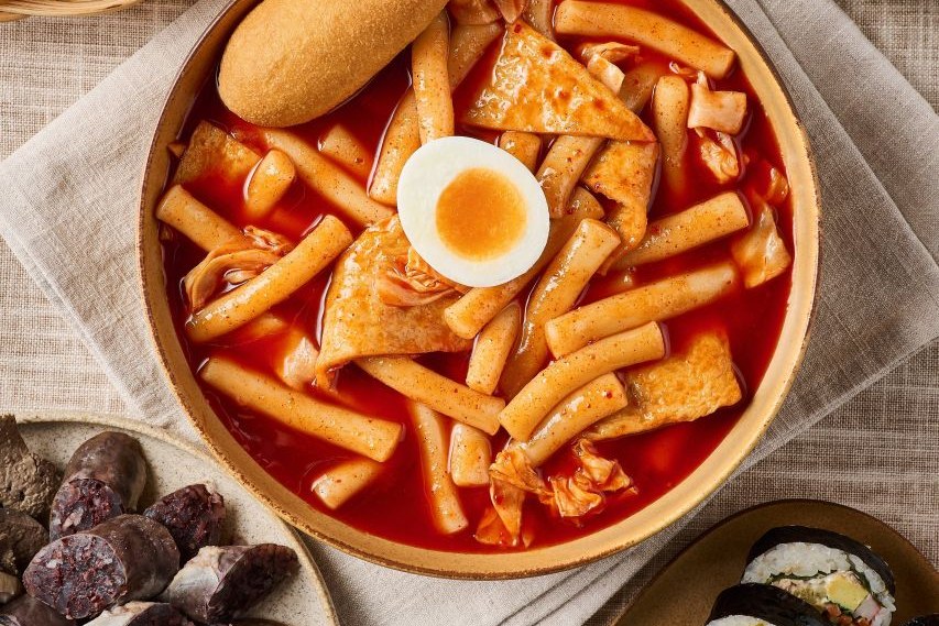 A bowl of tteokbokki, red rice cakes in sauce with a boiled egg half on top, seen from above, with part of a plate of blood sausage and gimbap at the edges
