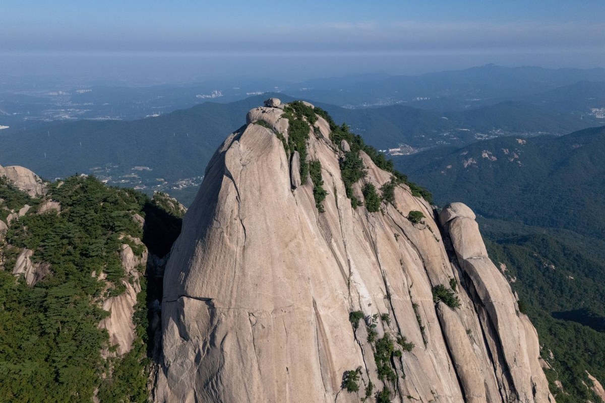 Aerial view of two rocky granite peaks of Bukhansan rising above green forest, with the city in haze behind