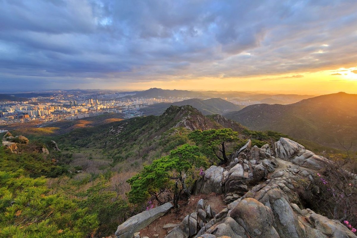 Sunset over Seoul seen from the rocky ridge of Gwanaksan, with trees in the foreground and a band of orange sky above the city