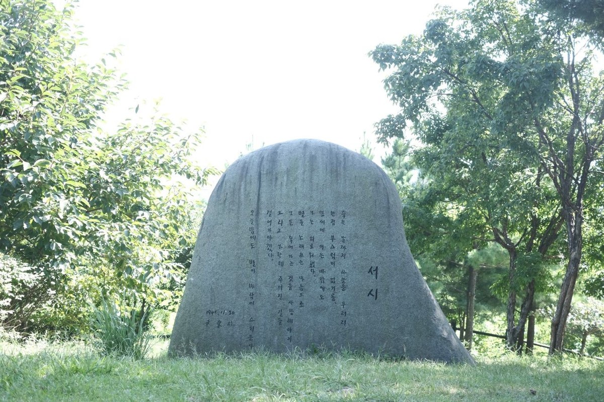 A large rounded stone carved with a poem in Korean standing among trees and grass at Poets' Hill