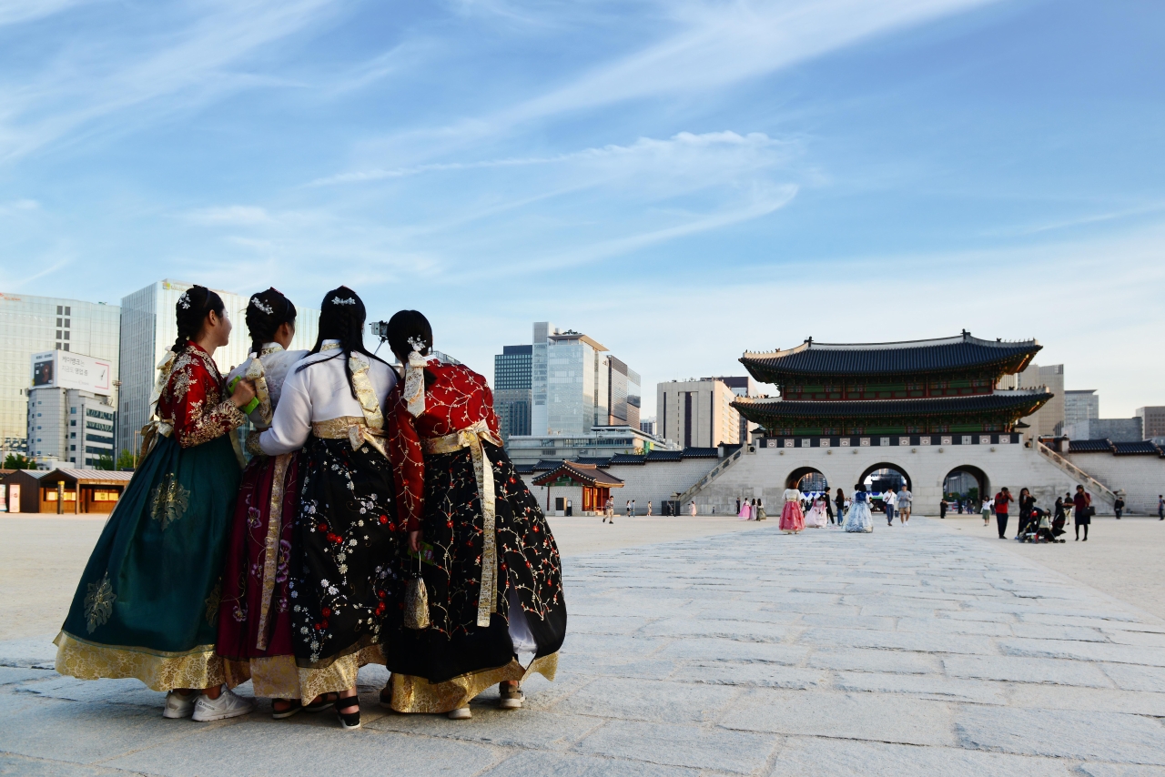Four visitors in colorful hanbok standing in the stone plaza in front of Gwanghwamun gate at Gyeongbokgung on a clear day