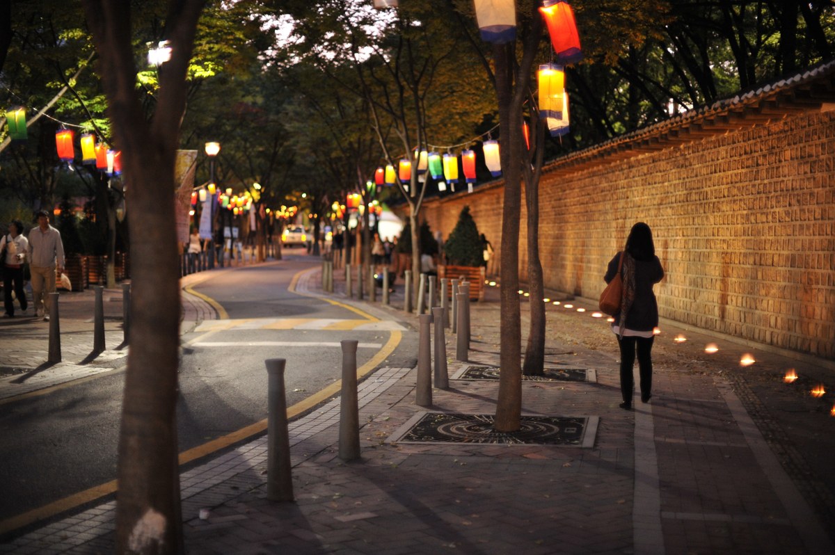 A woman walking along the brick-and-tile wall of Deoksugung at dusk, with colorful paper lanterns hanging from the trees above the road