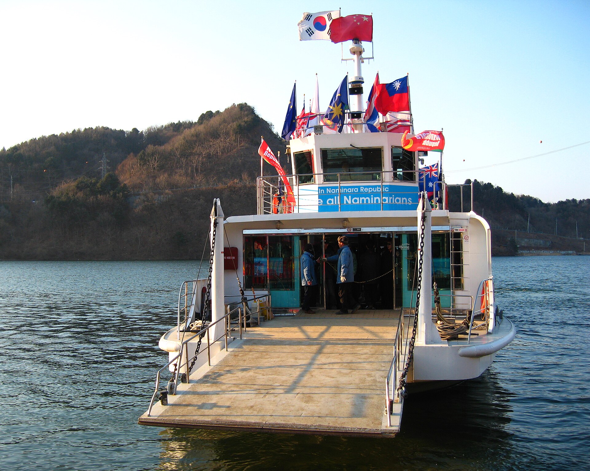 A white ferry with a ramp at the front on a calm lake, with flags along the top deck and forested hills behind