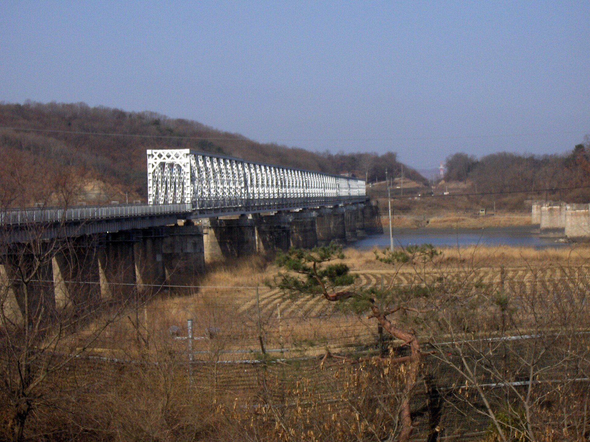 A steel bridge crossing a river toward bare hills under a pale sky, seen from a hillside near Imjingak Park