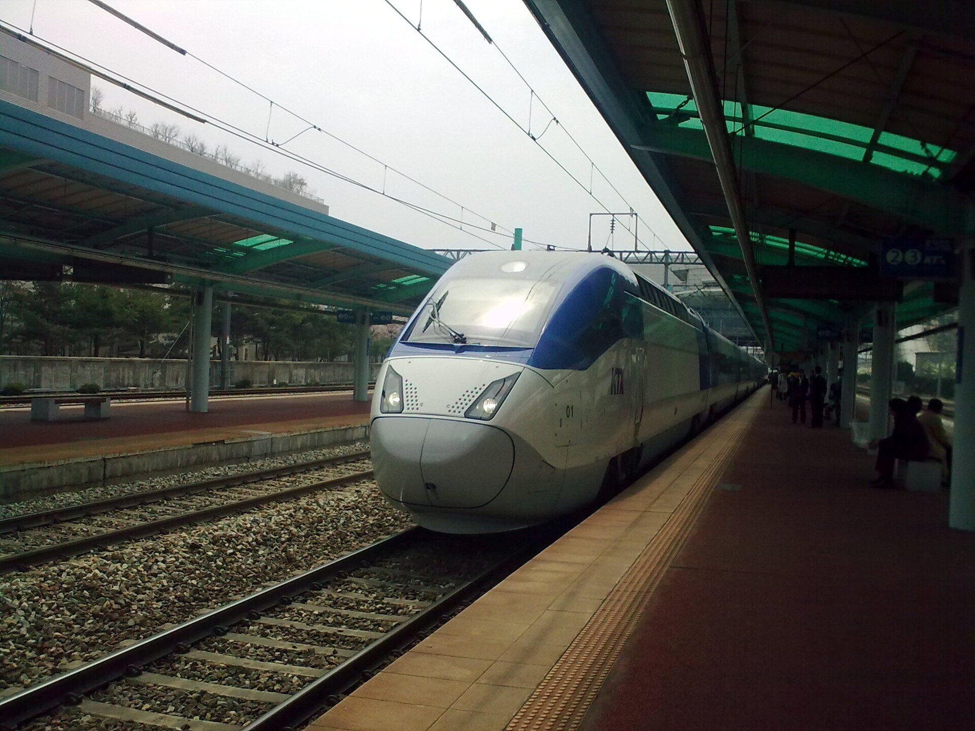 A white and blue KTX Sancheon high-speed train stopping at a platform with overhead power lines