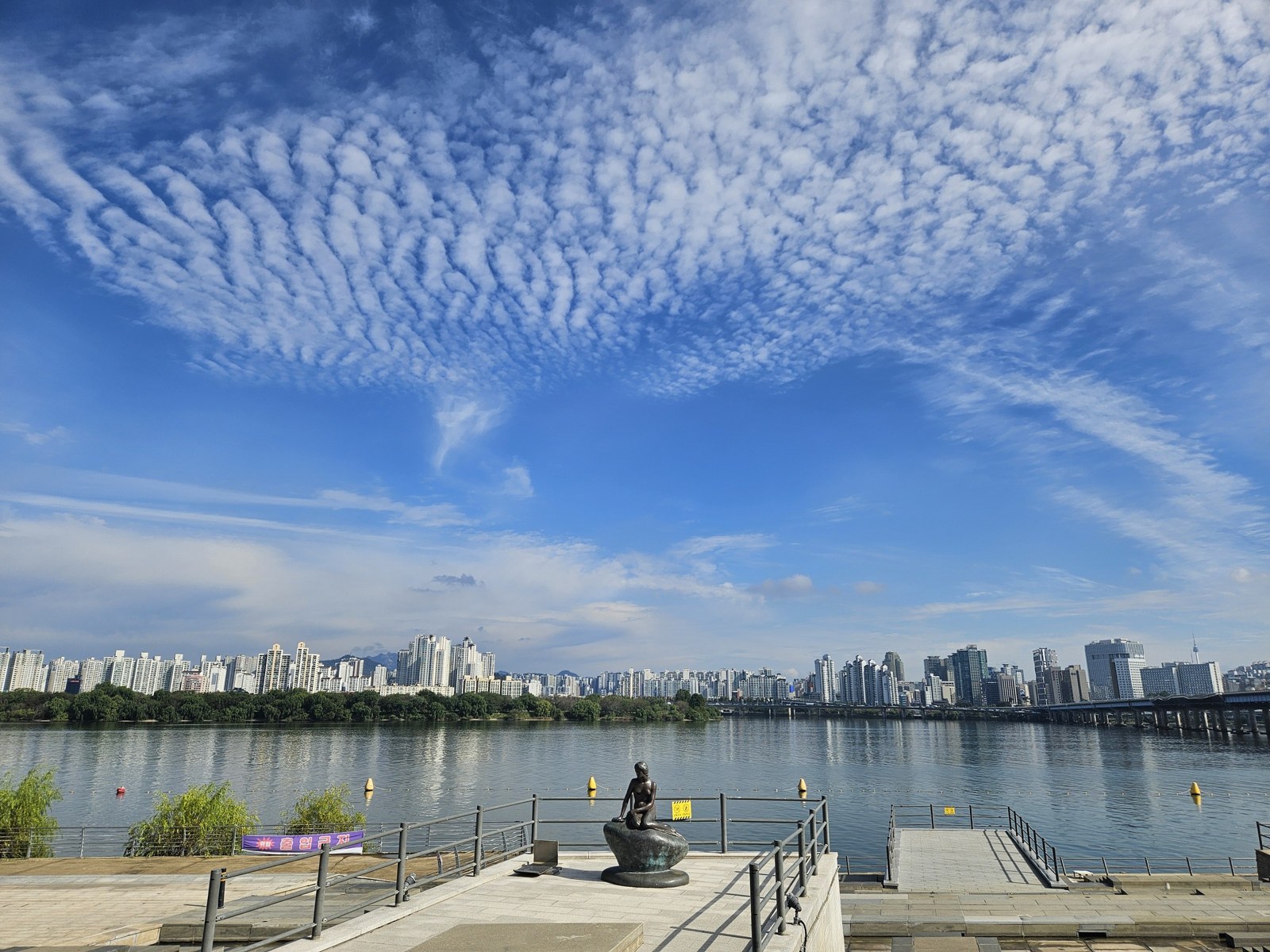 Han River with a bronze statue on a pier in the left foreground, the north-bank skyline and a sky full of altocumulus clouds