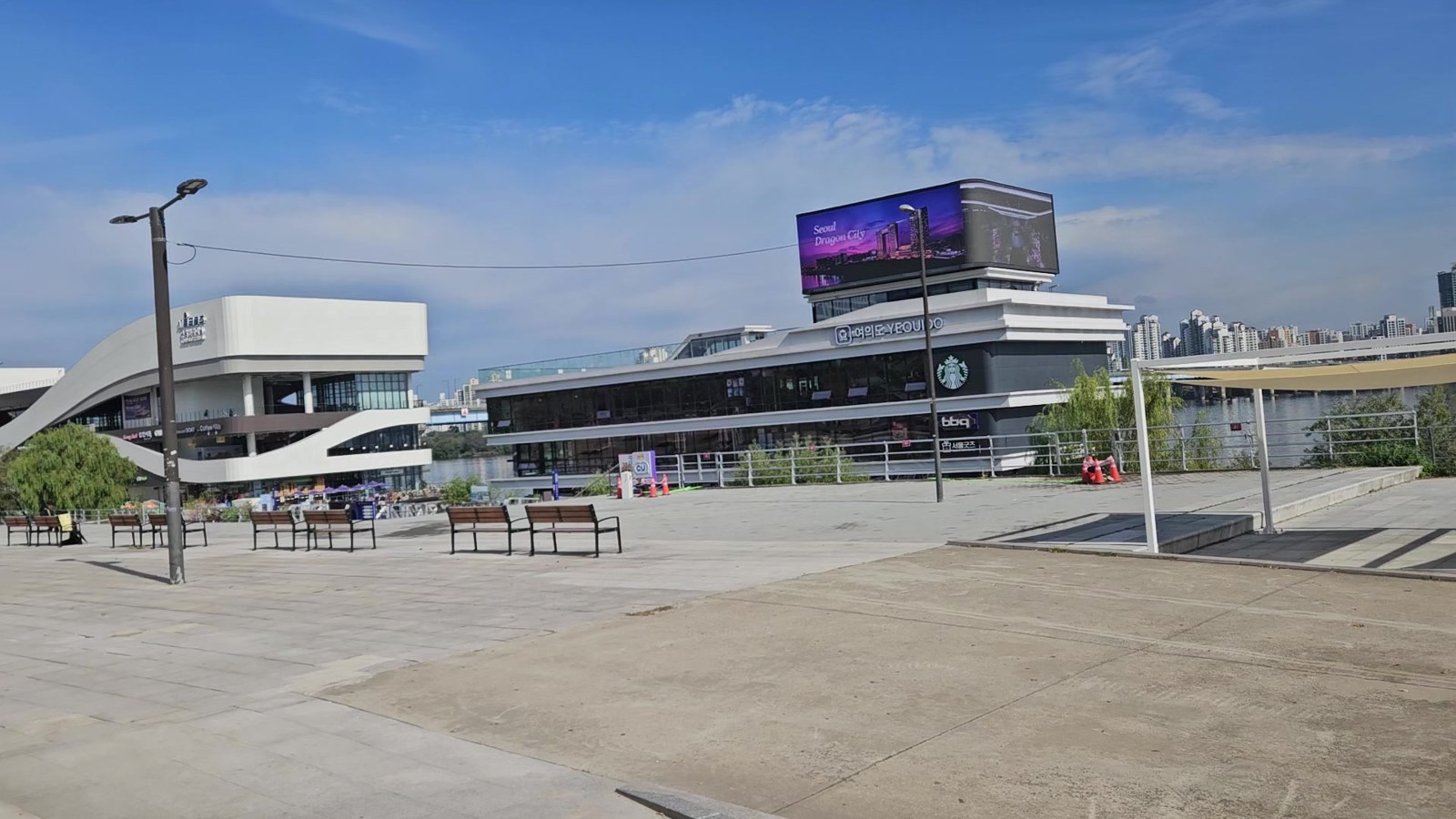 Wide paved plaza with benches under a blue sky, the white curved Seoul Cruise terminal on the left and the darker Han River Bus pier building with a roof screen and Starbucks sign on the right