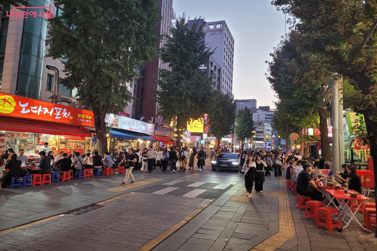 Crowded pojangmacha street at dusk in Jongno 3-ga, Seoul, with red plastic stools and tables lining a pedestrian lane under ginkgo trees and shop signs lighting up
