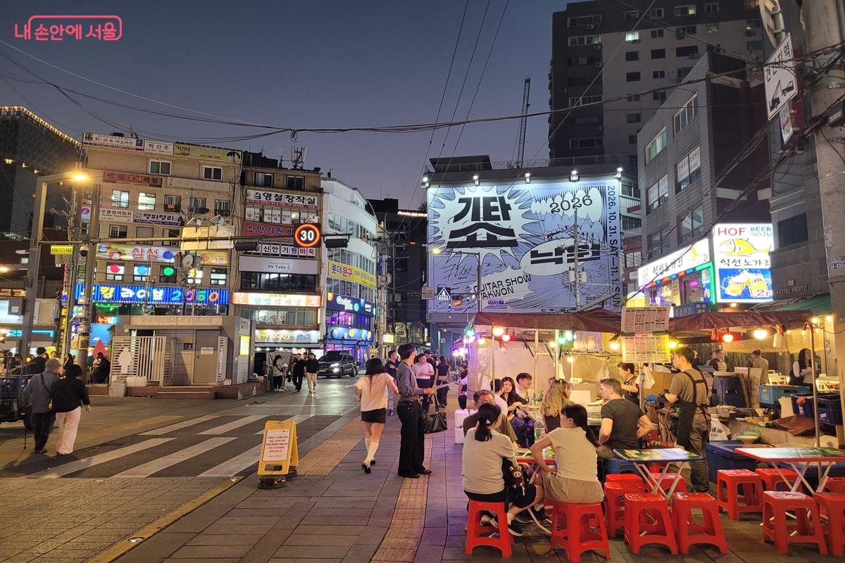 Outdoor tables with red stools in front of lit-up pojangmacha tents at a Jongno 3-ga street corner at night, with a crosswalk and neon signs behind