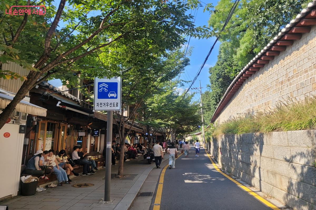 Paved lane beside the long Jongmyo stone wall in Seoul with hanok cafes and people sitting outside under trees