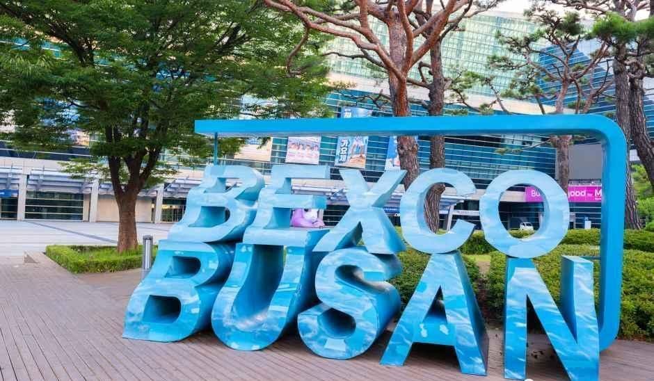 The blue BEXCO BUSAN letter sign on the plaza outside BEXCO in Haeundae, Busan, with trees and glass buildings behind