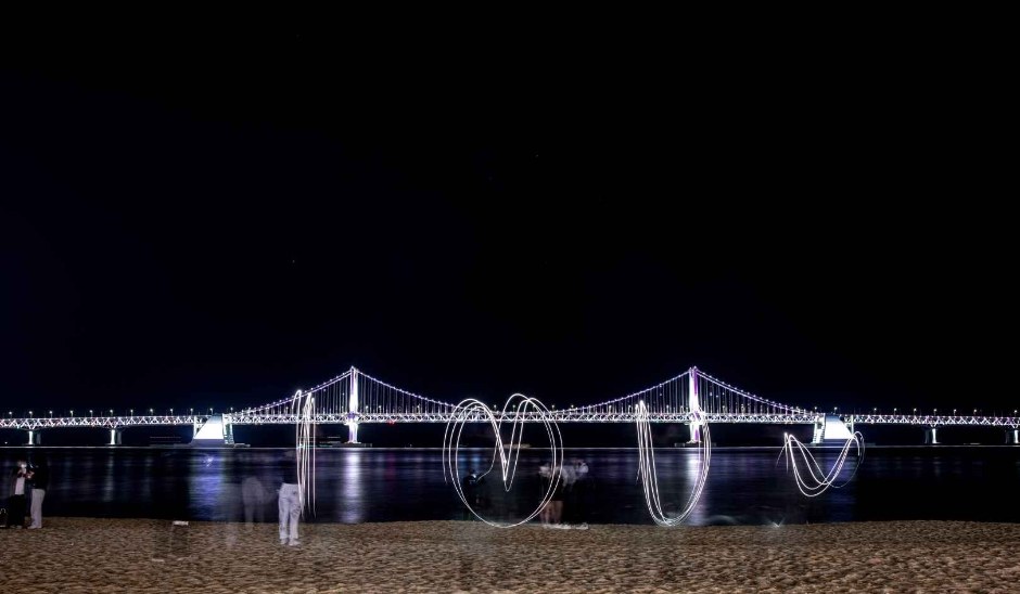 Gwangan Bridge lit up at night over the sand at Gwangalli Beach in Busan, with white light trails drawn on the beach in a long exposure