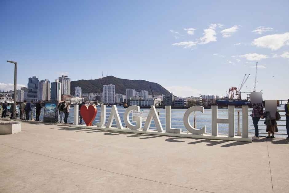 The white I love JAGALCHI sign with a red heart on the waterfront plaza at Jagalchi Market in Busan, with the harbor and a hill behind