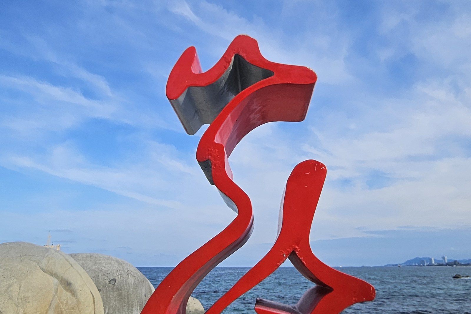 A bright red abstract sculpture on a black base with round boulders and waves of the sea behind it
