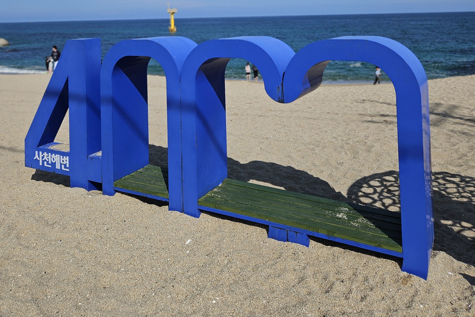A large blue sculpture on the sand spelling out the beach name in Korean, with the sea and a yellow marker tower behind it
