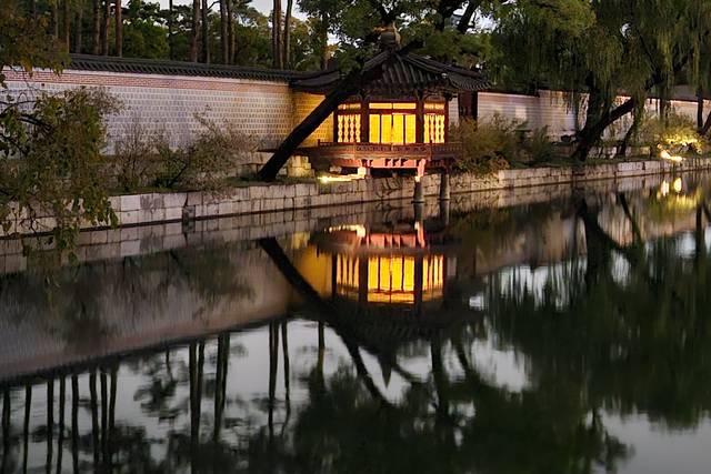 A lit wooden pavilion with willow trees reflected in a still pond inside a Seoul palace at dusk